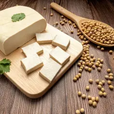 Blok jasnego tofu, pokrojony na mniejsze kawałki, leży na desce. Obok znajduje się drewniana łyżka z ziarnami soi, pokazująca, jak wygląda tofu.