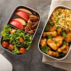 Dwa pojemniki z jedzeniem: jeden z kurczakiem w sosie, ryżem i natką, drugi z sałatką, jabłkiem i orzechami. Idealne lunchbox do pracy przepisy.