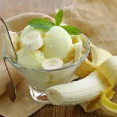 Pyszne lody z mrożonych bananów, udekorowane plasterkami banana i listkiem mięty, w szklanej miseczce. Obok leży obrany banan.