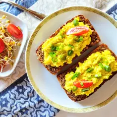 Pyszne, żółte tofu na ciemnym pieczywie, posypane szczypiorkiem i pomidorkami. Idealne tofu na śniadanie, obok miseczka kiełków z pomidorkami.