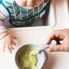 Maluch w śliniaczku je awokado. Rodzic podaje mu łyżeczkę z zielonym puree. Proste przepisy na awokado dla niemowlaka.