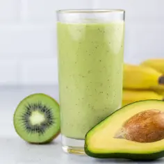 Zielony koktajl z awokado, kiwi i bananów w szklance, obok połówka awokado i kiwi.