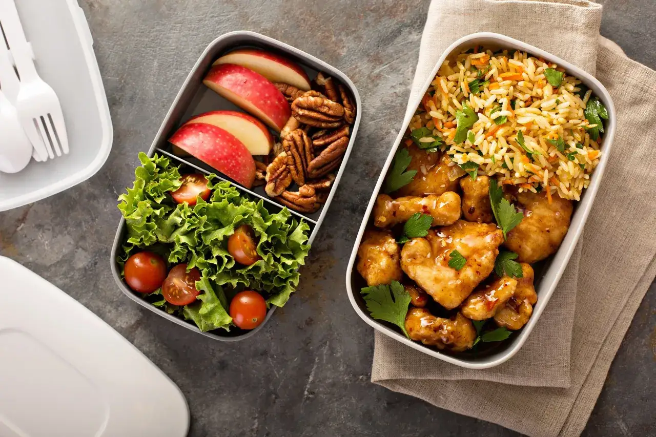 Dwa pojemniki z jedzeniem: jeden z kurczakiem w sosie, ryżem i natką, drugi z sałatką, pomidorkami, jabłkiem i orzechami. Inspiracja na lunchbox do pracy przepisy.