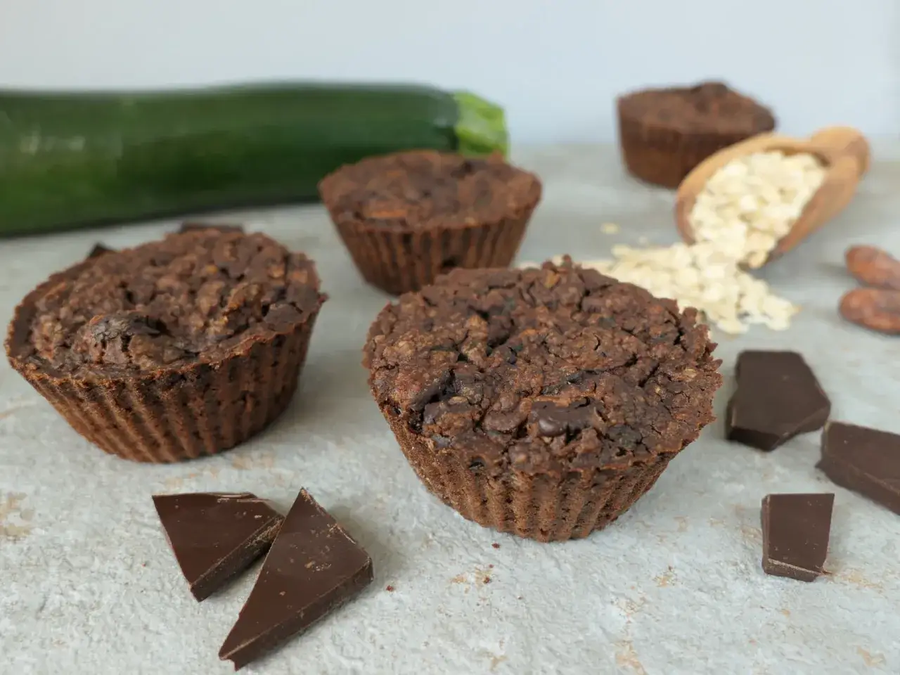 Czekoladowe muffinki z cukinią, obok kawałki czekolady, ziarna kakaowca i płatki owsiane.
