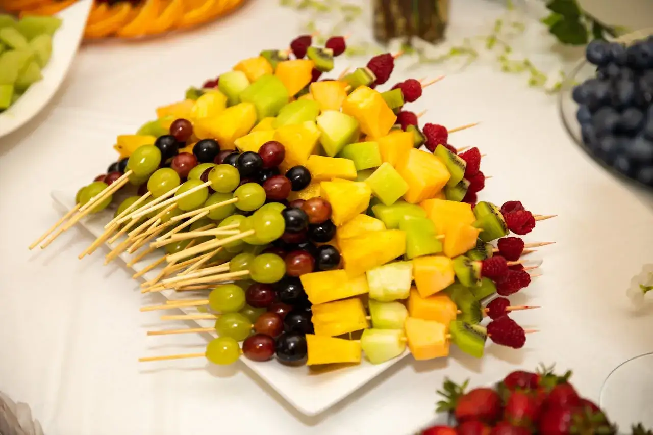 Kolorowe szaszłyki owocowe na talerzu – idealny sposób, jak efektownie podać owoce na przyjęciu. Winogrona, kiwi, mango i maliny.