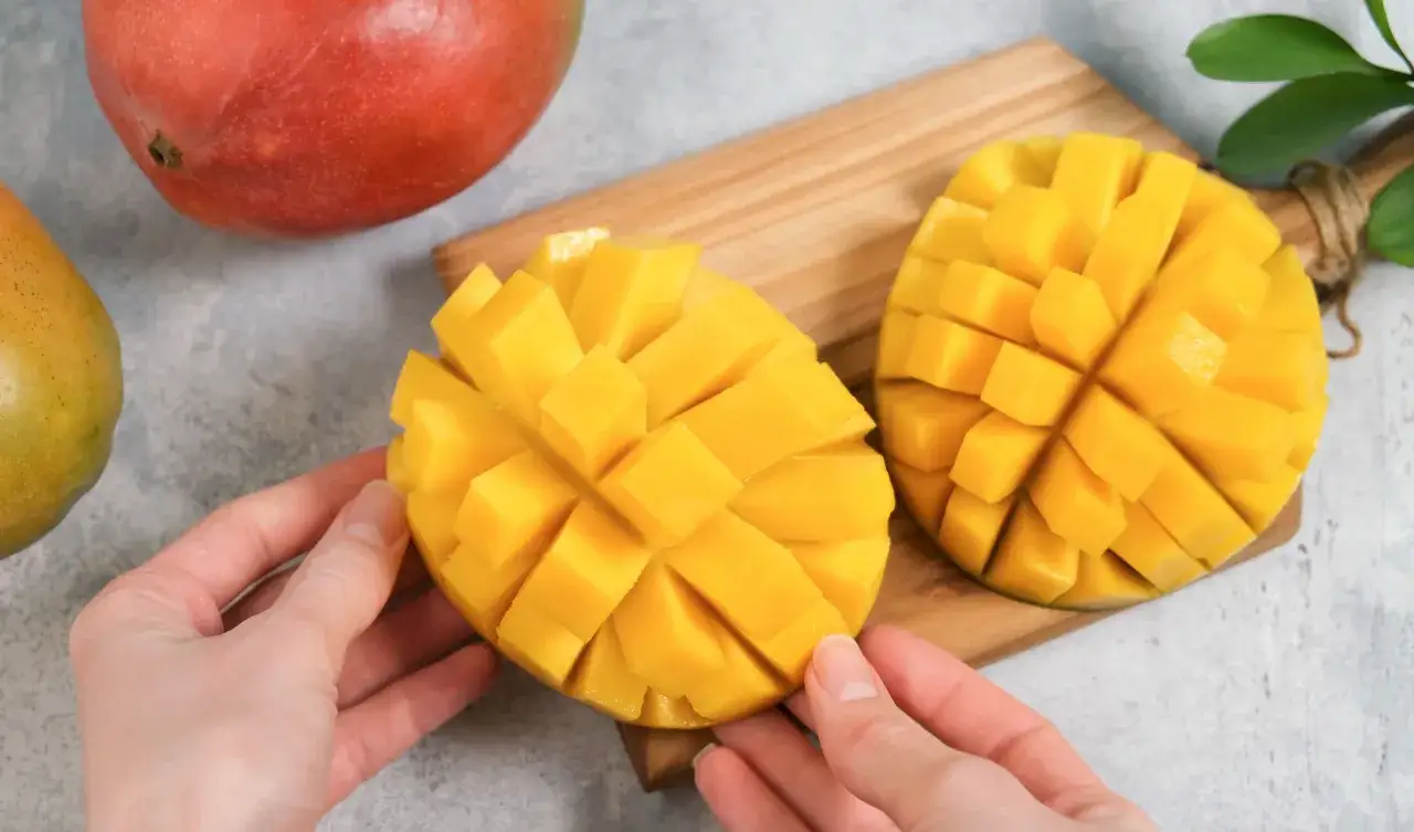Dwa pokrojone w kostkę mango na desce, gotowe do jedzenia. Obok całe, czerwone mango.