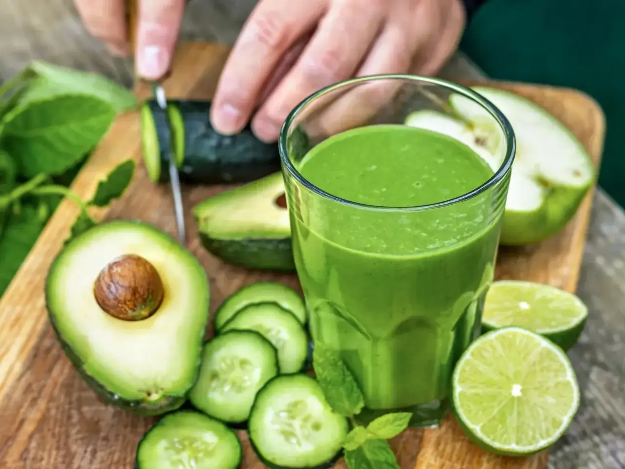 Zielony koktajl z awokado, ogórka i limonki na drewnianej desce. Obok szklanki leżą połówki awokado, plasterki ogórka i limonki.