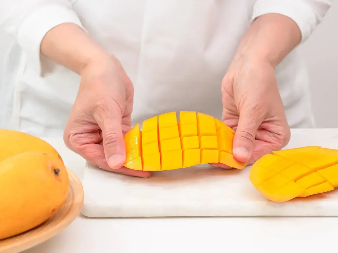 Ręce kroją mango owoc jak jeść, tworząc kosteczki. Obok leżą całe owoce.