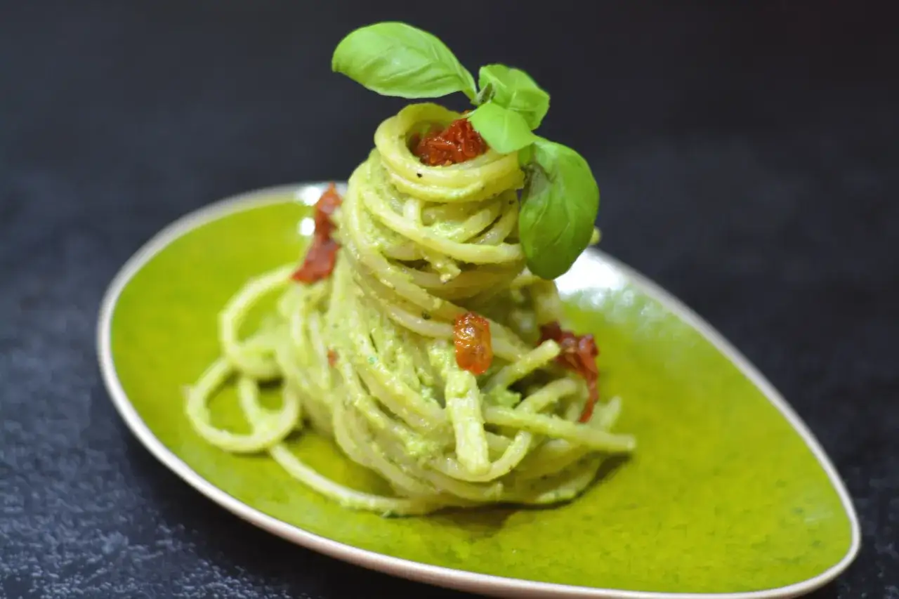 Spaghetti z kremowym pesto z awokado, ozdobione suszonymi pomidorami i listkami bazylii, na zielonym talerzu.