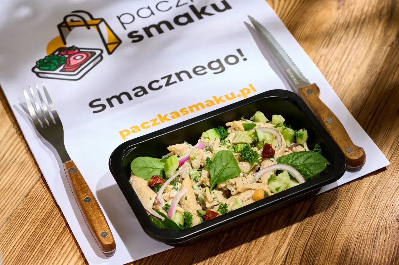 Pyszna dieta pudełkowa w Nowym Sączu. Sałatka z kurczakiem, warzywami i makaronem w czarnym pojemniku, obok widelec i nóż.