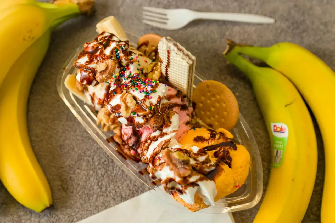 Pyszne lody z mrożonych bananów, polane czekoladą, posypane orzechami i kolorową posypką. Obok leżą świeże banany.