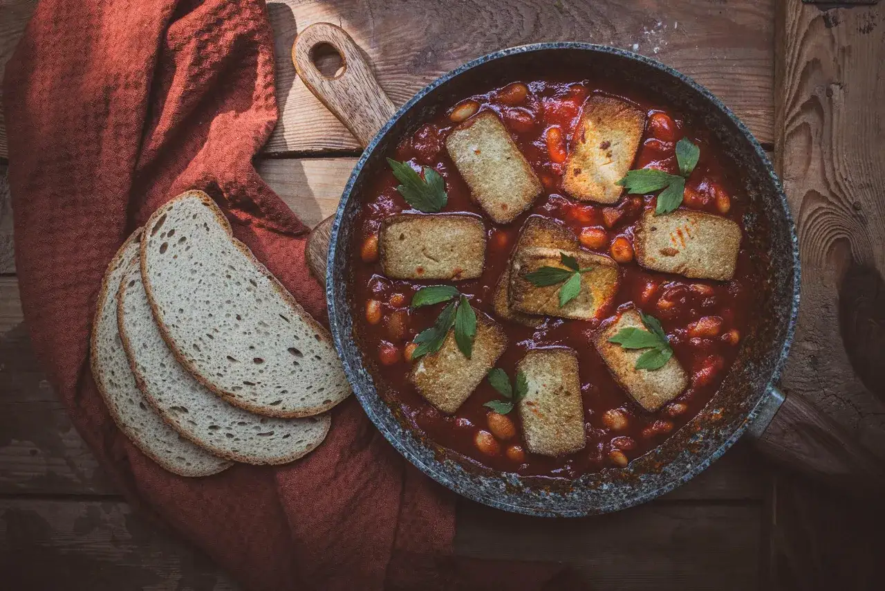 Smażone tofu w sosie pomidorowym z fasolką, podane w patelni, obok kromek chleba.