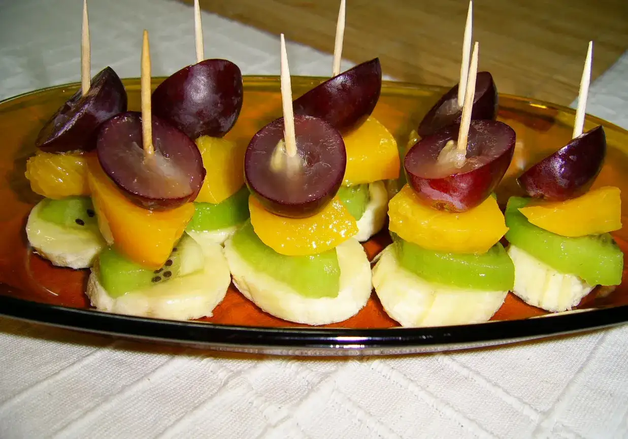 Kolorowe owoce na patyczkach: banan, kiwi, brzoskwinia, winogrono i pomarańcza – idealne co do koreczków na przyjęcie.