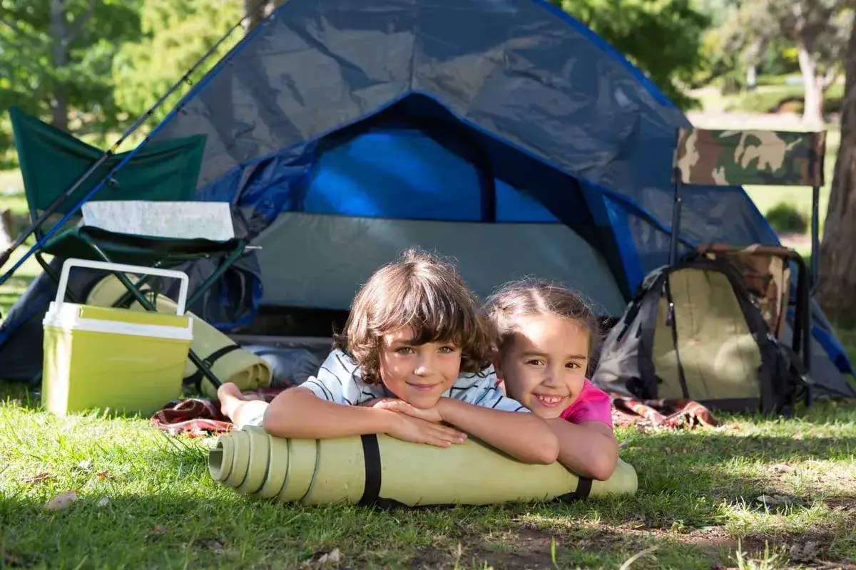 Dzieci leżą na macie przed namiotem, gotowe na przygodę. Pomyśl, co zabrać pod namiot z dziećmi: lodówka, śpiwory, krzesła.
