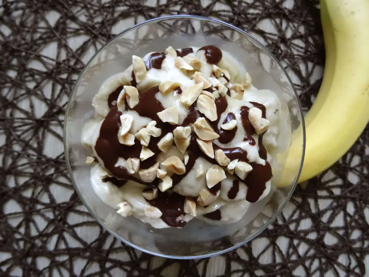 Pyszne lody z mrożonych bananów polane czekoladą i posypane orzeszkami. Obok leży świeży banan.